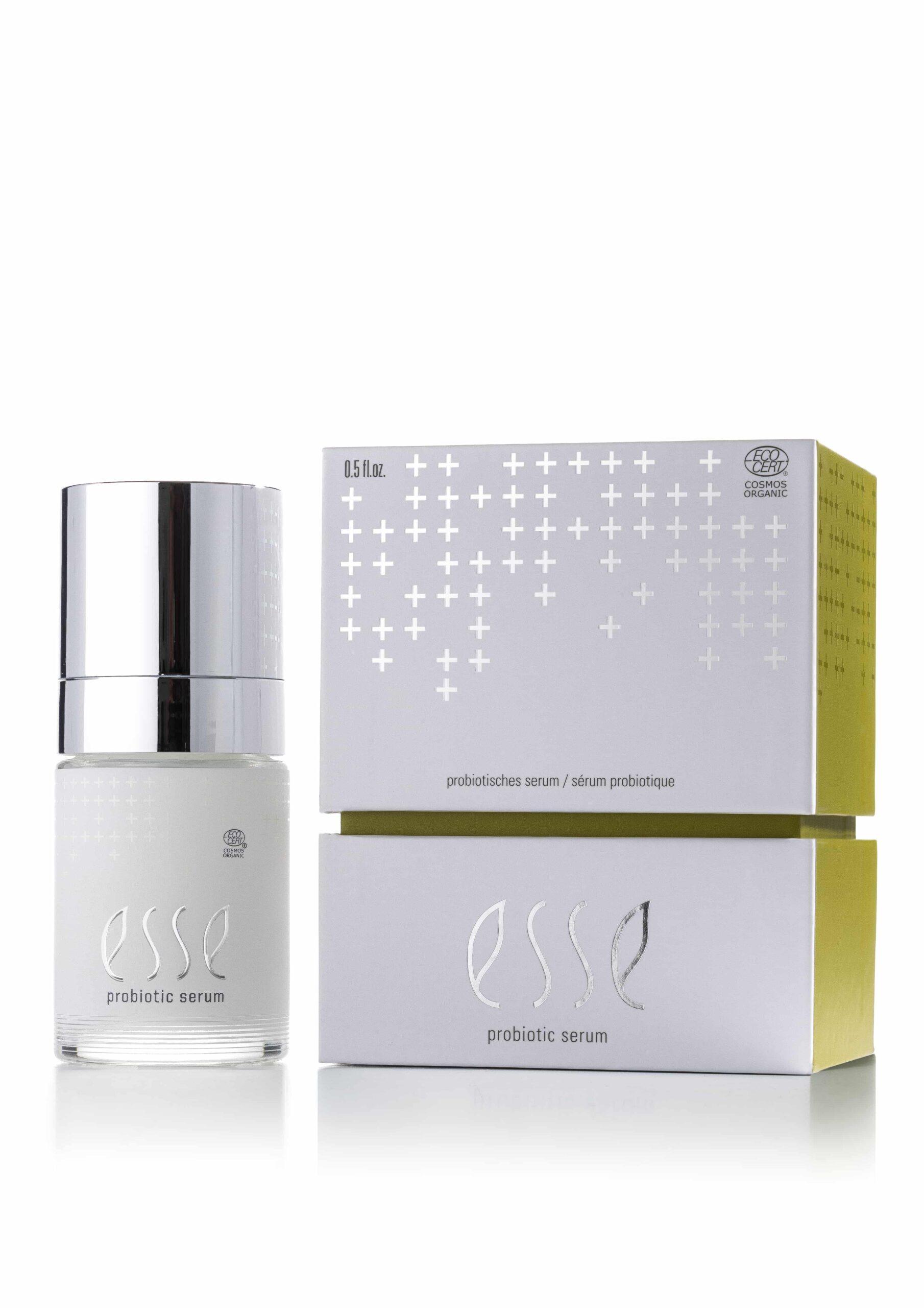 En flaska Esse Probiotic Serum med silverlock står bredvid den vitgula förpackningen med silverfärgad text och ett plusteckensmönster.