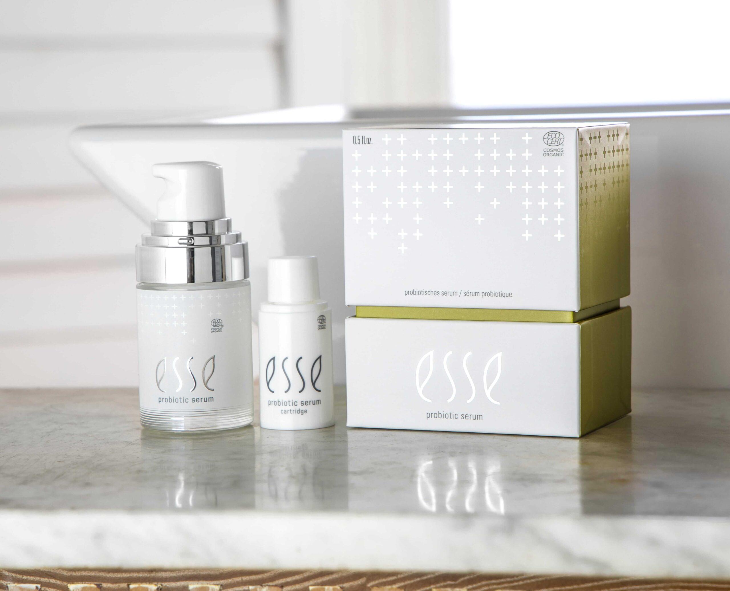 Två flaskor med Esse Probiotic Serum - en pump och en liten flaska - står på en marmoryta bredvid två staplade märkeslådor, en vit och en grön.